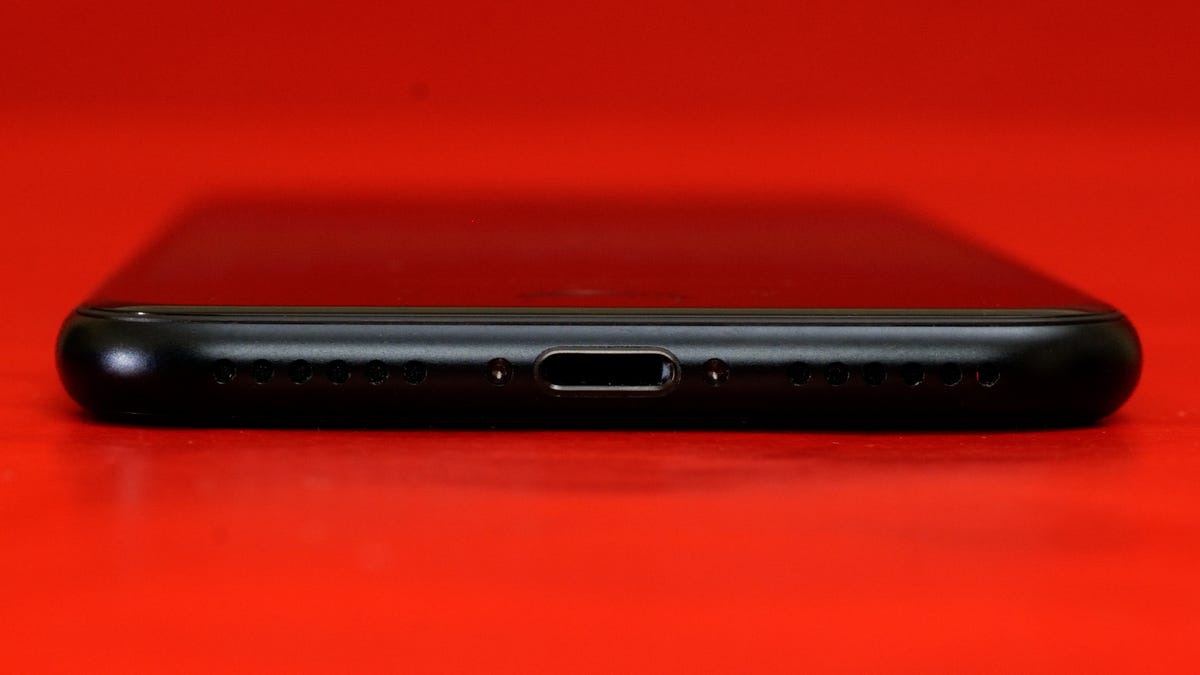 A close up shot of the iPhone SE 2022 charging port, the black phone is laying flat on a red background. 