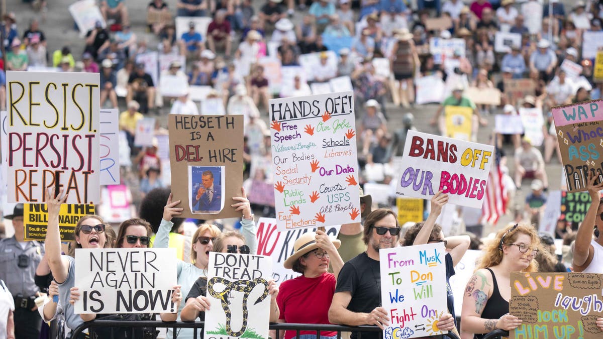 A large crowd of protesters holds up handmade signs at a public demonstration. Visible signs include slogans like 