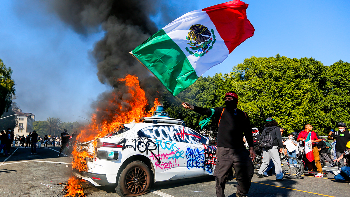 A protester waves the Mexican flag in front of a burning Waymo vehicle during an anti-ICE protest in downtown Los Angeles on June 8, 2025.
