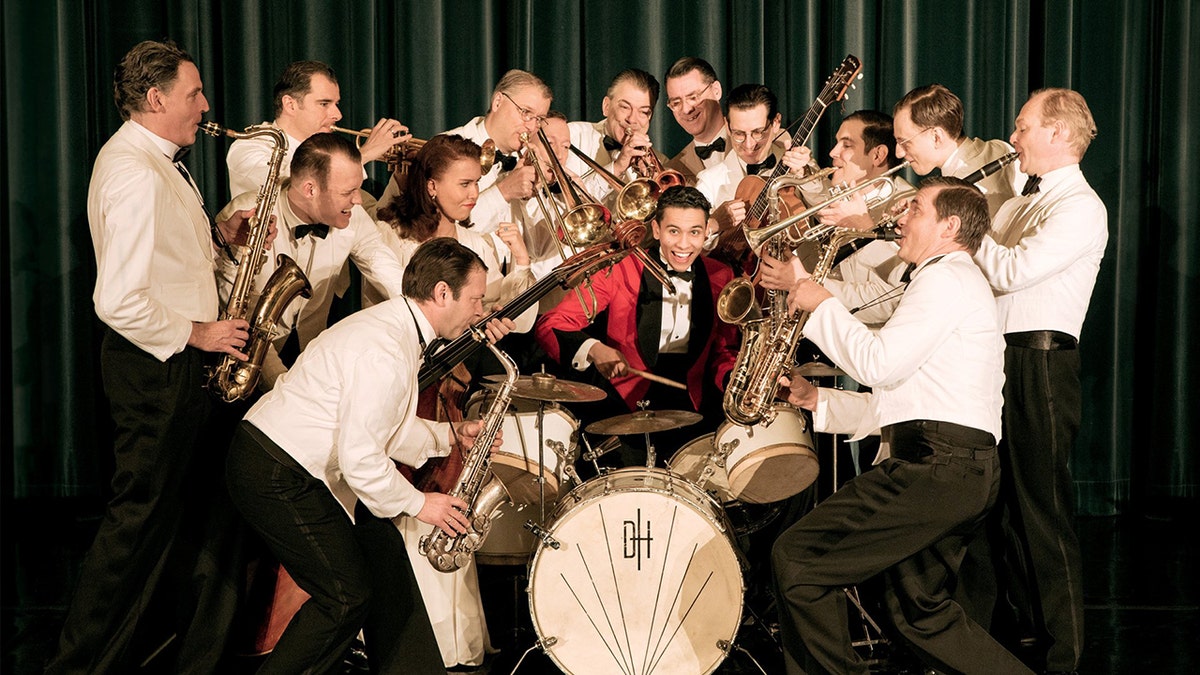 David Hermlin on drums with large swing orchestra playing brass instruments around him.