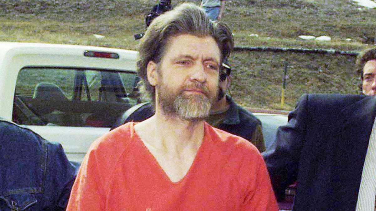 Ted Kaczynski walks alongside law enforcement officers wearing an orange jumpsuit