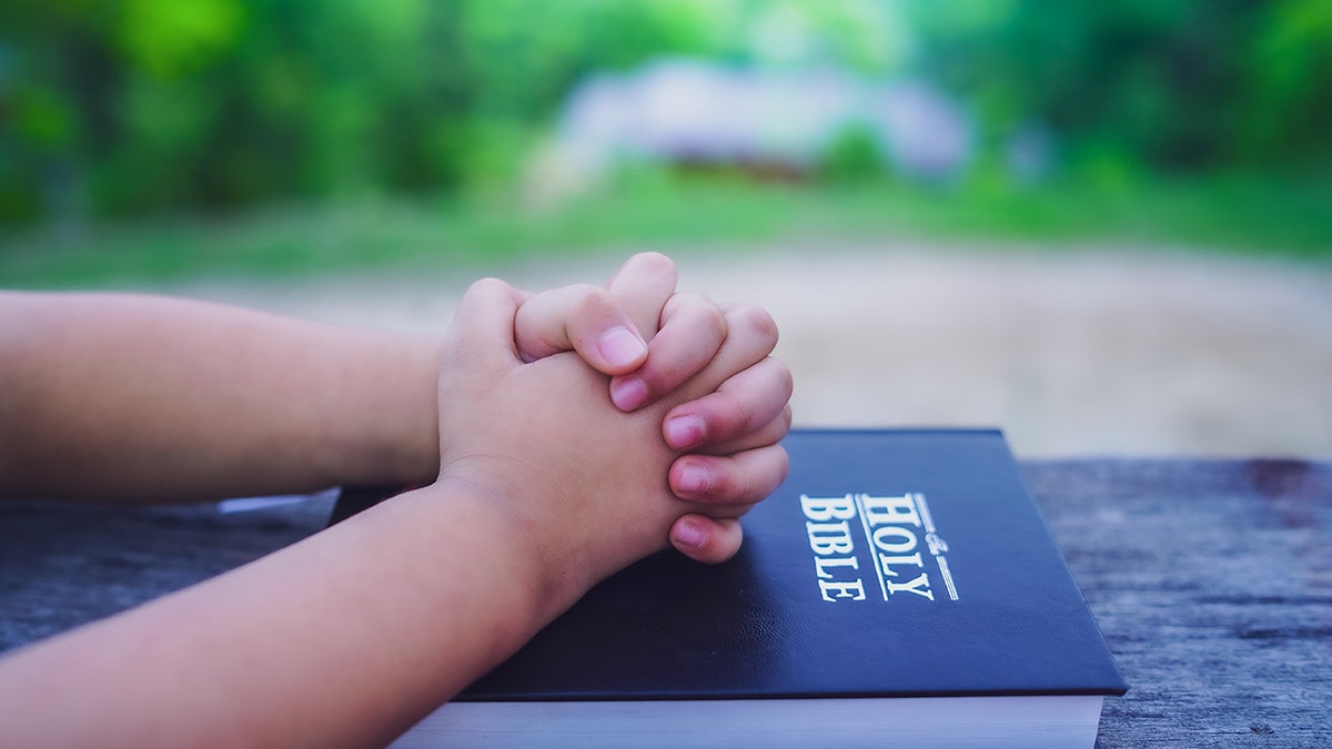 Close-up of a child’s hands folded in prayer resting on a Bible