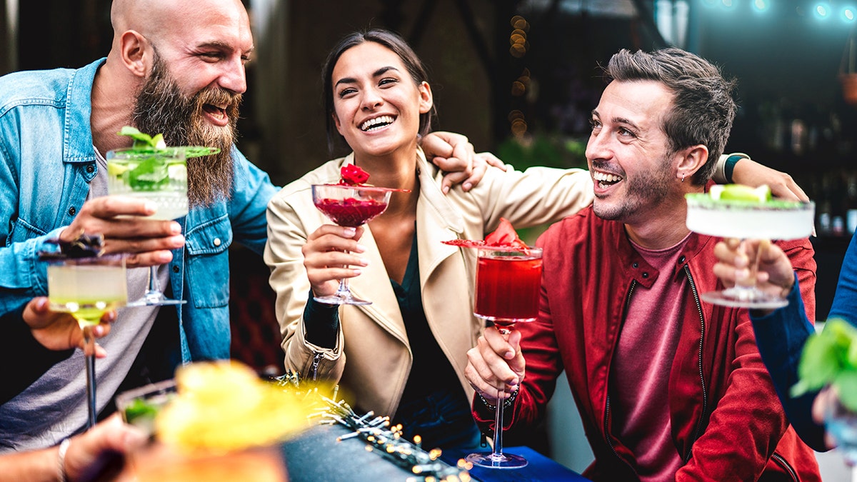 Friends laughing and toasting with colorful mocktails at an outdoor bar.