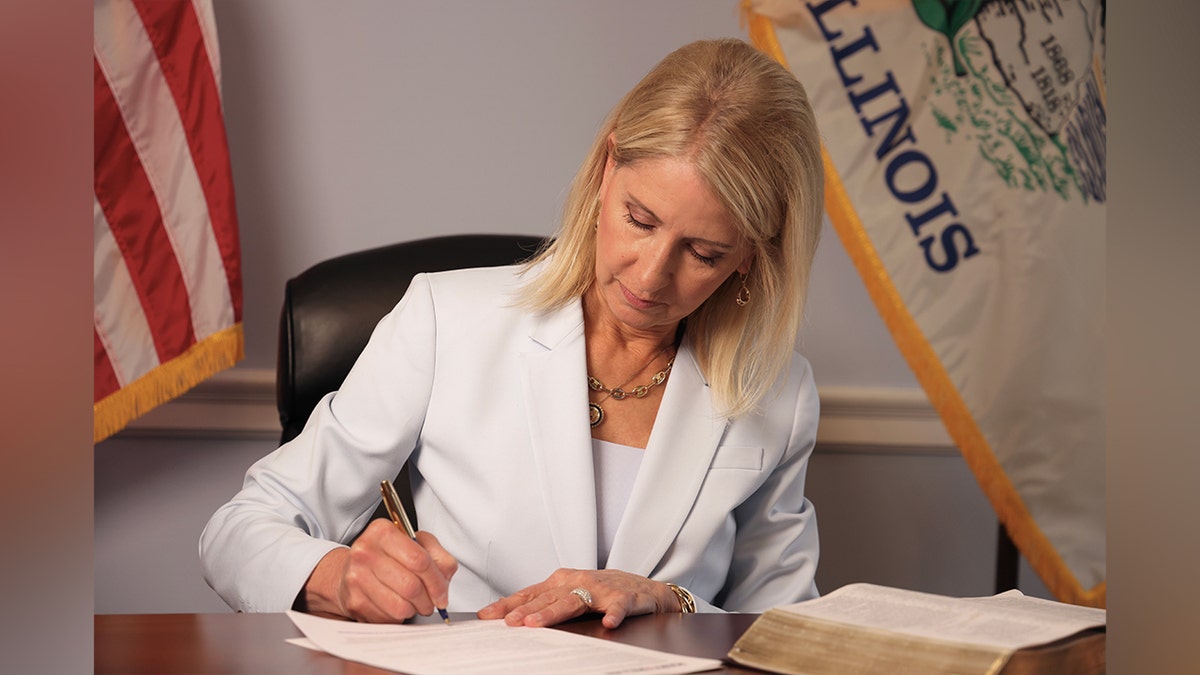 Rep. Mary Miller signs paperwork at her desk with state and national flags behind her.
