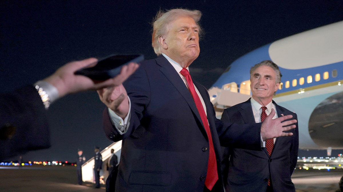 Trump speaks to journalists on an airport tarmac while a cabinet official stands nearby.