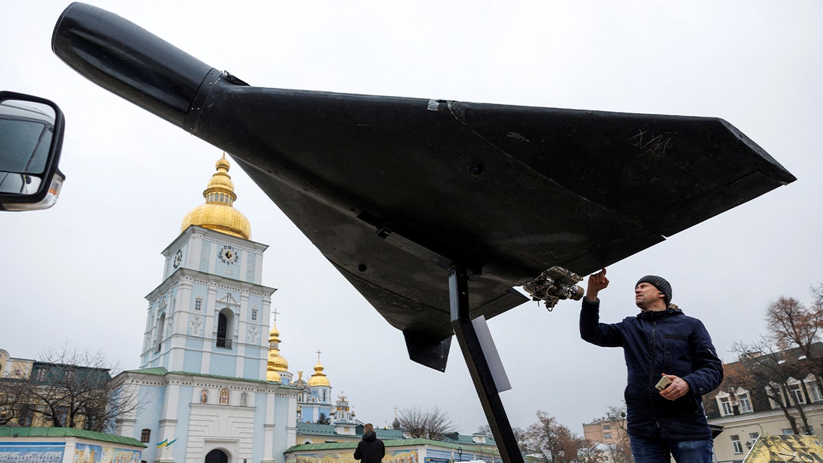 A person reaches out to touch a displayed Shahed-136 drone set up outdoors near a cathedral.