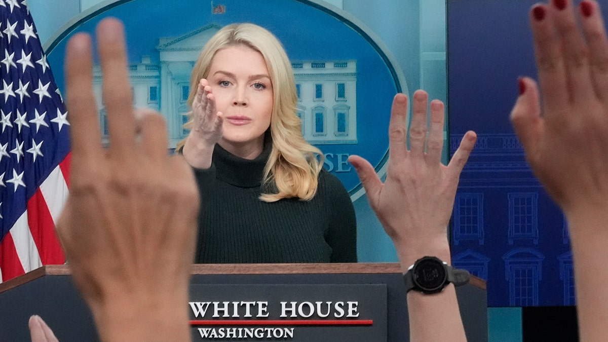 White House Press Secretary Karoline Leavitt speaking at a podium while reporters raise their hands.