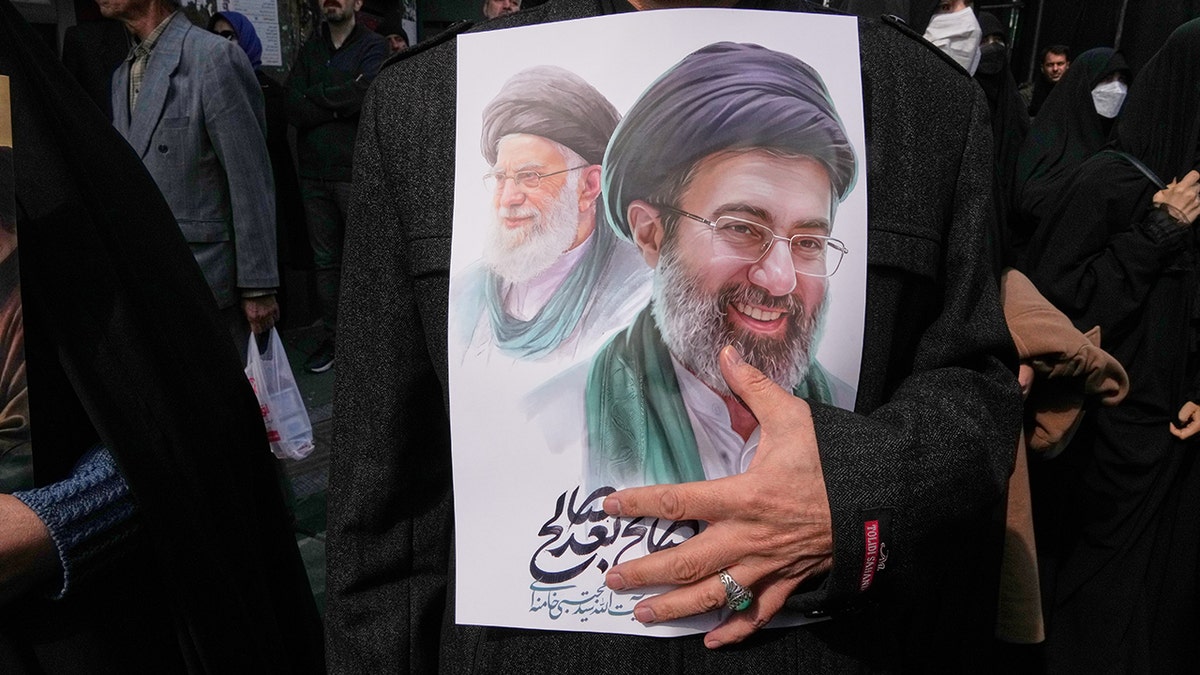 A person holding a poster depicting Ayatollah Mojtaba Khamenei and Ayatollah Ali Khamenei during a funeral procession.