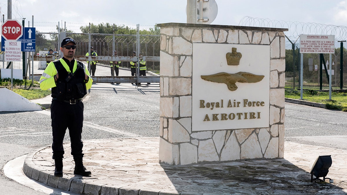 A police officer stands next to a sign that reads "Royal Air Force AKROTIRI" near a gated road.