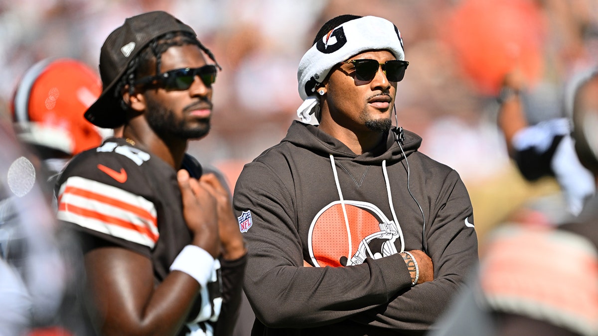 Quarterbacks Shedeur Sanders and Deshaun Watson standing on the sidelines during a football game.