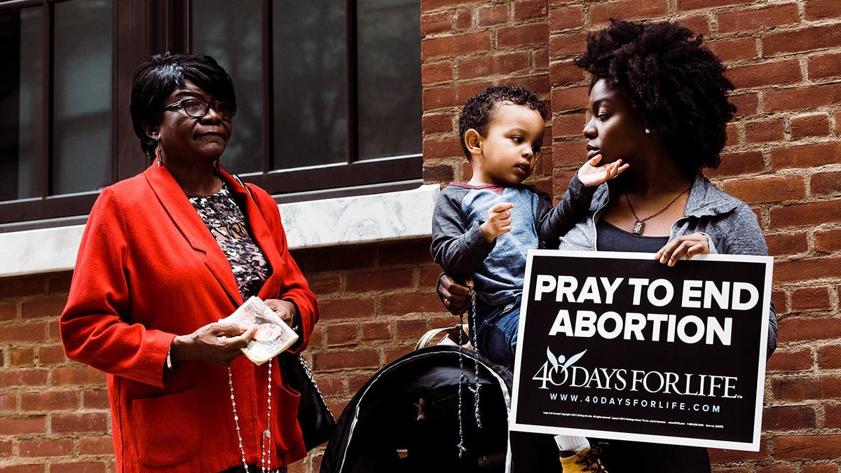 Two women and a child stand outside a building, with one holding a sign that reads 