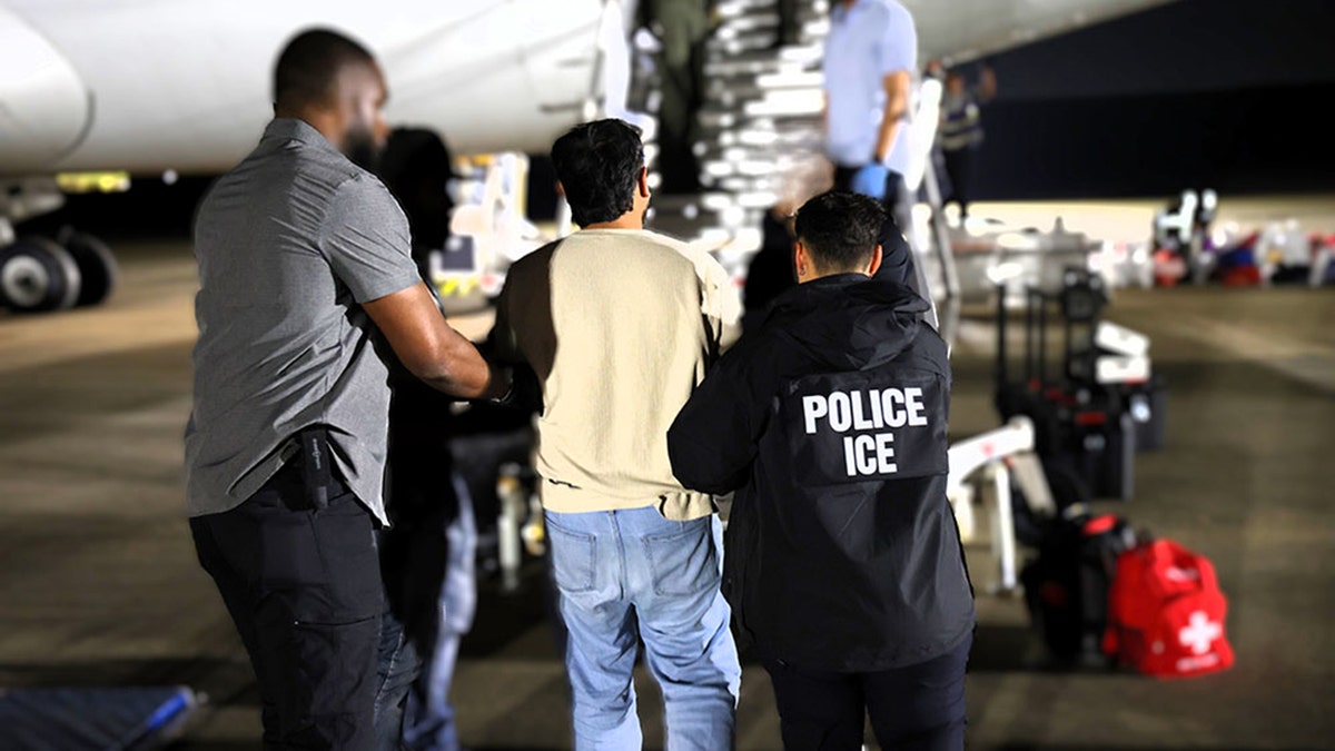 ICE personnel escort a male detainee in hand restraints up stairs to board a deportation plane