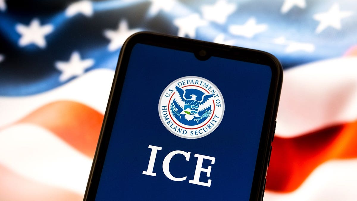 Smartphone displaying U.S. Department of Homeland Security and ICE emblem with U.S. flag on laptop screen behind