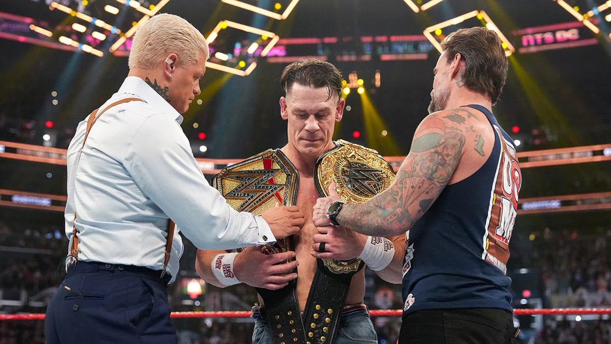 Cody Rhodes and CM Punk presenting John Cena with WWE championship belts in arena