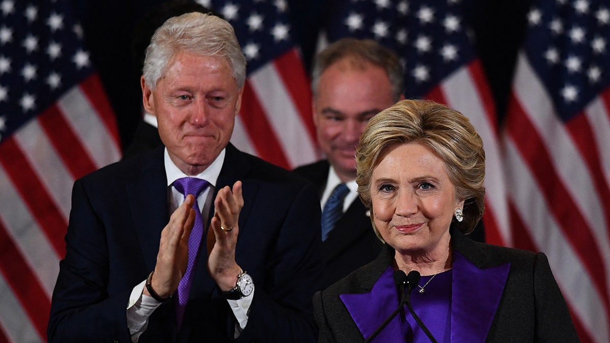Hillary Clinton delivering a concession speech with Bill Clinton and Tim Kaine seated behind her in New York