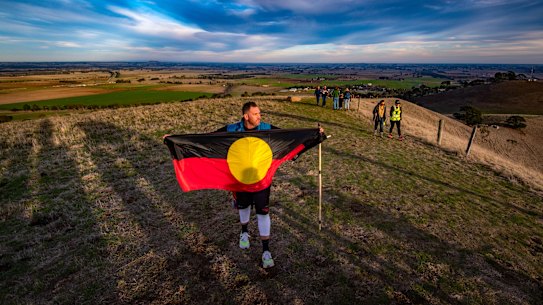 Travis Lovett unfurls the Aboriginal flag at the top of Mount Noorat during his Yoorrook Justice Commission Walk for Truth. The site was an important meeting place for First Nations people of western Victoria.