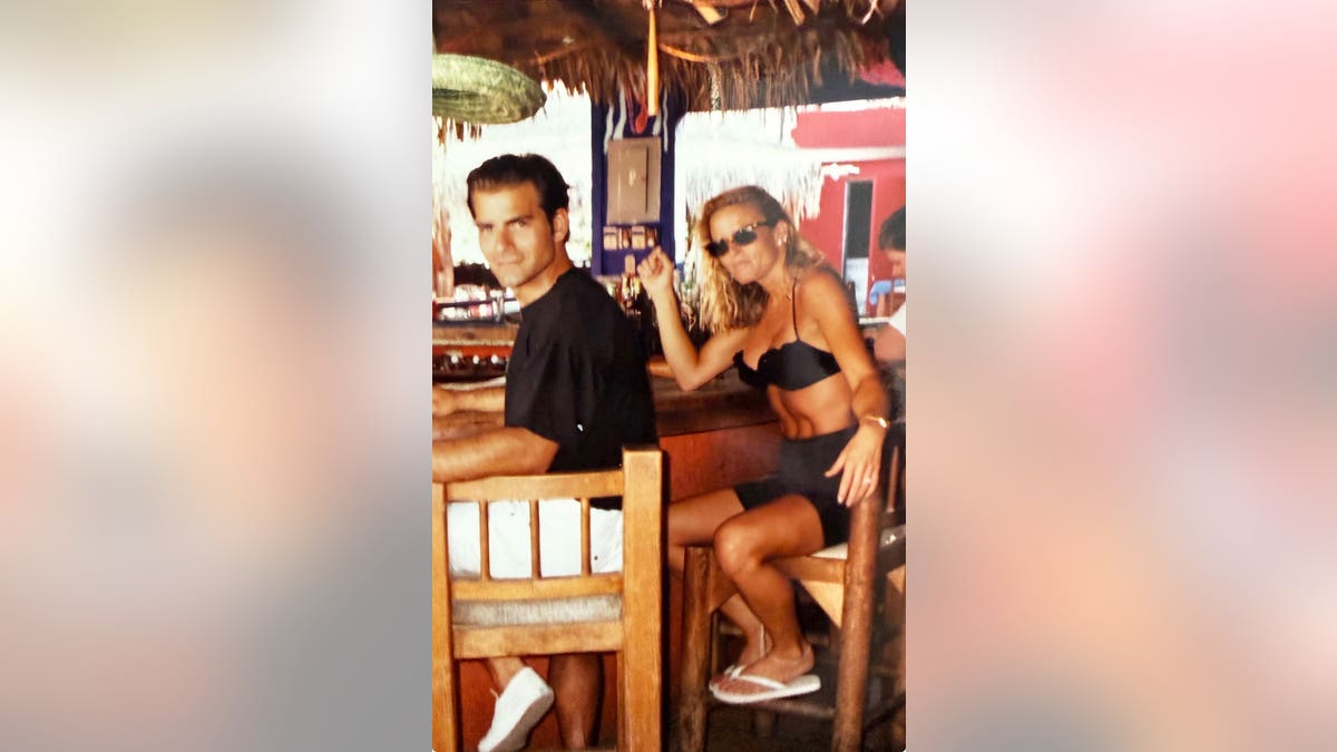 Nicole Brown Simpson in a black bikini sitting next to Joseph Perrulli in a black shirt.