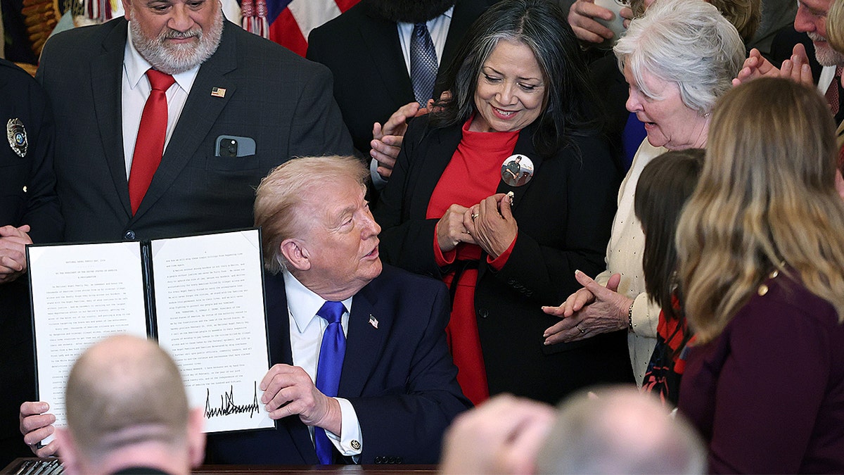 Donald Trump holding up a signed proclamation paper while standing in the East Room of the White House.