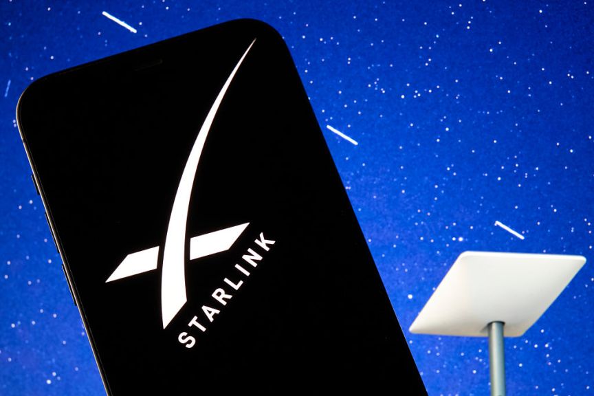 The SpaceX Starlink logo is seen displayed on a smartphone screen, with a Starlink satellite dish visible in the background.