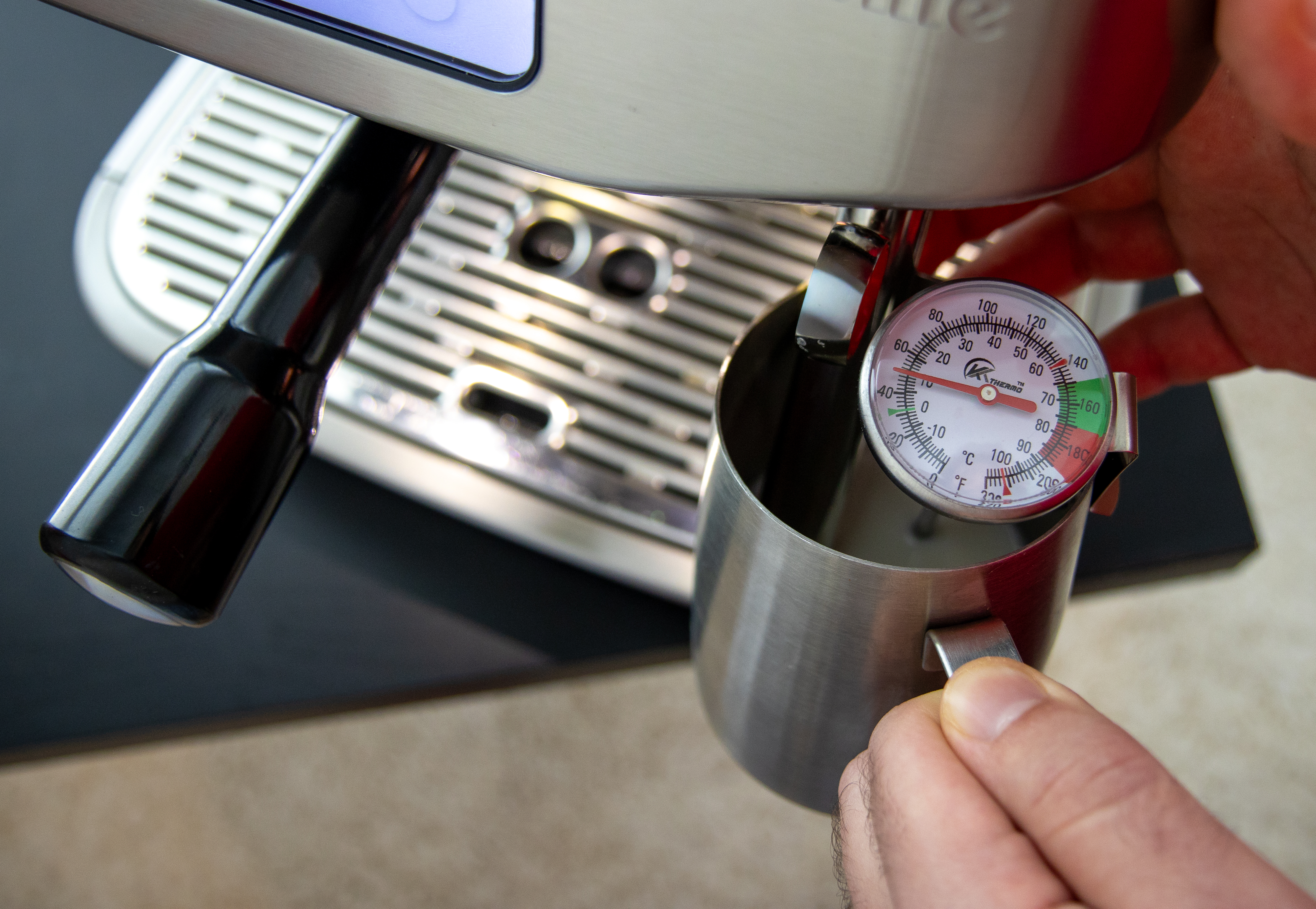 Milk is being steamed using an espresso maker with an instant-read thermometer clipped to a metal milk pitcher.