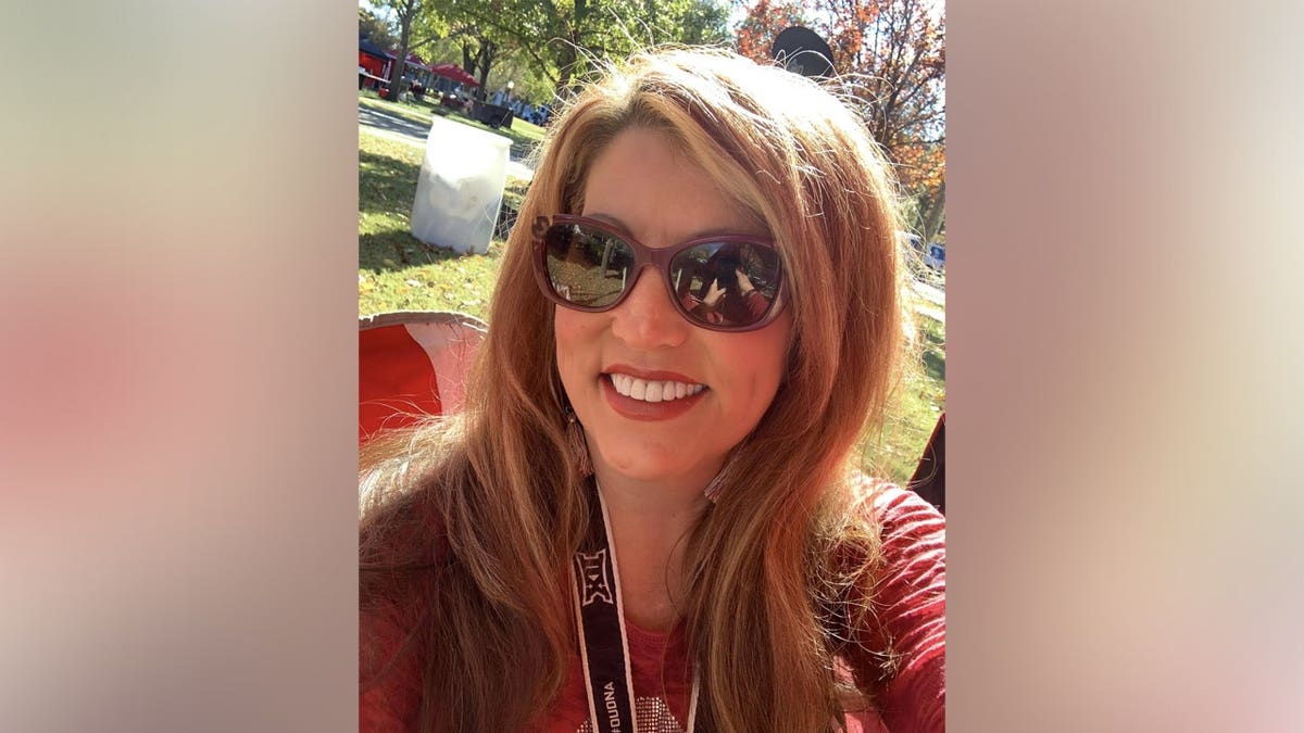 Jenny Lupton smiles for a selfie outdoors on a sunny day, wearing sunglasses and a lanyard.
