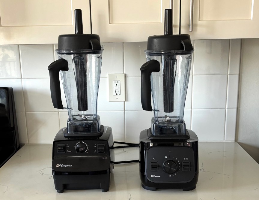 The Vitamix 5200 blender on the left and the VX1 on the right, both in black, on a white marble kitchen countertop.