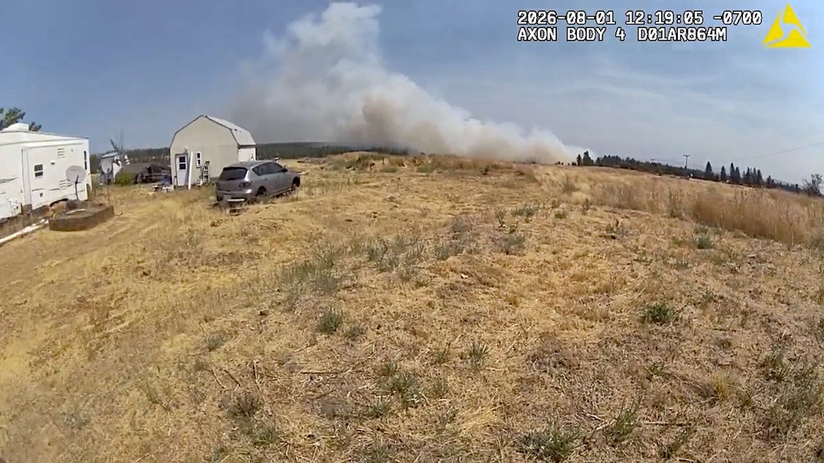 Thick smoke rises beyond farm equipment, a trailer and a small shed in a dry rural area.
