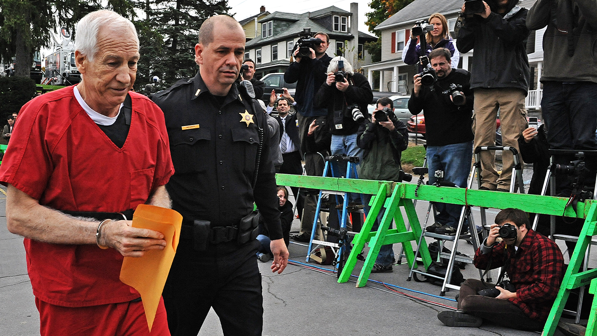 Jerry Sandusky in handcuffs and a jail jumpsuit carries a large envelop into court for his sentencing