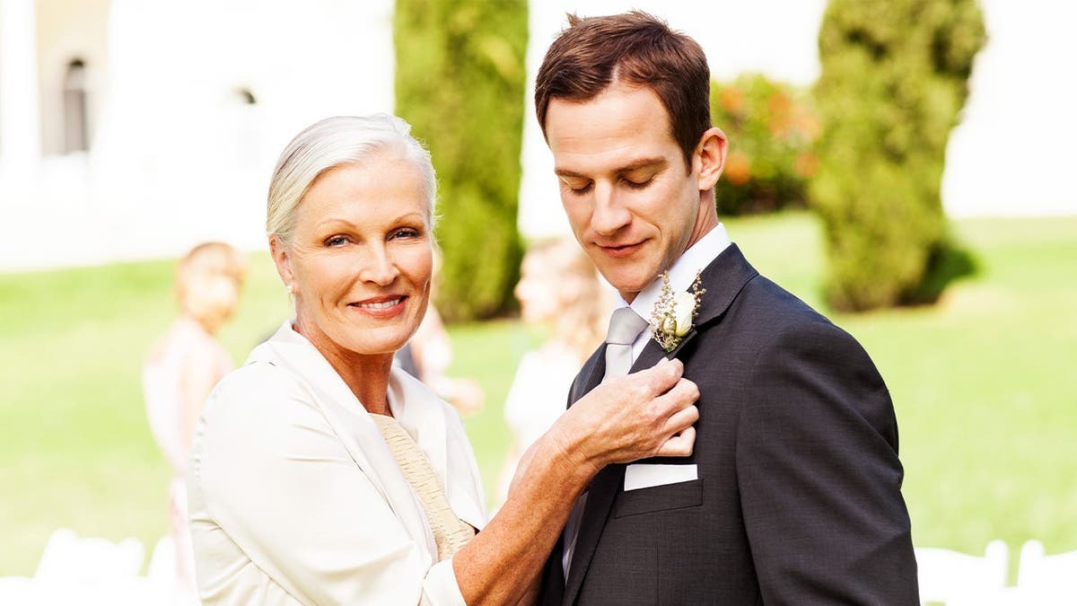 A mother-of-the-groom wearing white pins a flower to the groom's jacket.