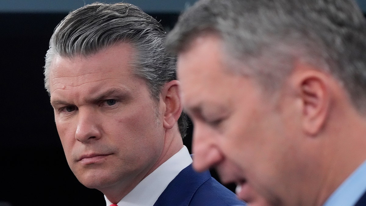 Pete Hegseth looking toward the camera while Gen. Dan Caine speaks in the foreground during a briefing.
